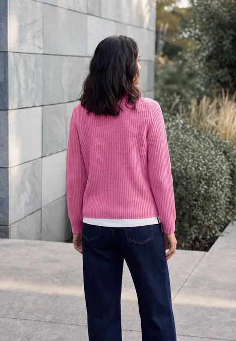Trui met rits pink flash melange Frau in pinkem Pullover und dunkler Jeans steht im Freien, Rücken zur Kamera, vor Mauer und Büschen.