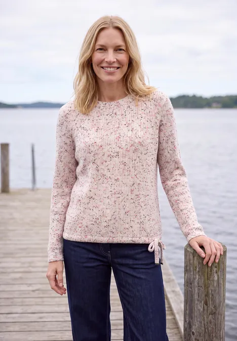 Eine lächelnde Frau in Pullover und Jeans steht entspannt an einem Holzsteg am See.