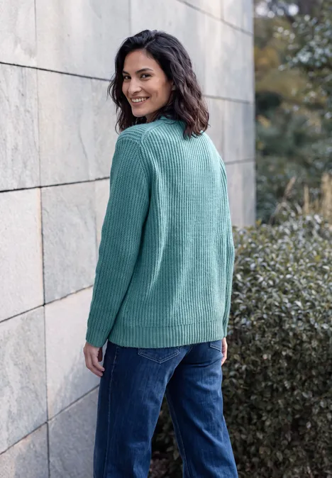 Een vrouw met krullend haar draagt een groene trui en een blauwe jeans, glimlachend tegen een stenen muur