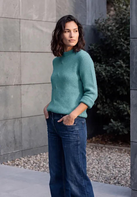 Een vrouw in een turquoise trui en jeans leunt ontspannen met handen in de zakken tegen een grijze muur.