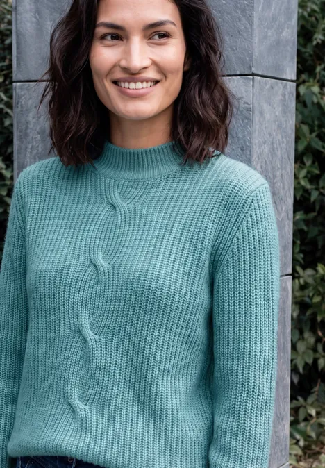 Een glimlachende vrouw in een mintgroene trui voor een grijze stenen muur en planten.