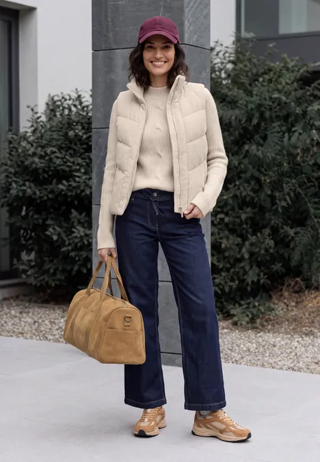 Trui met col en vlechtmotief oat milk beige melange Vrouw met paarse pet, beige bodywarmer, trui, wijde jeans, bruine schoenen en tas voor een gebouw.