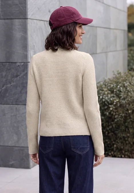 Trui met col en vlechtmotief oat milk beige melange Een vrouw met een crèmekleurige trui, donkere jeans en een bordeauxrood petje, gezien van achteren buiten.