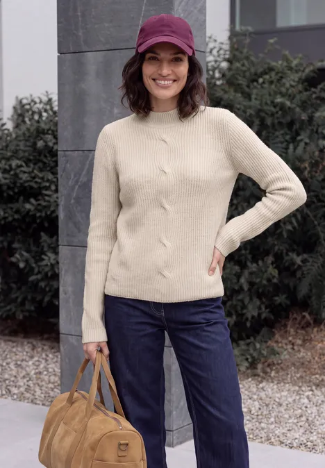 Trui met col en vlechtmotief oat milk beige melange Vrouw in beige trui, blauwe jeans en paarse pet, met een bruine tas, glimlachend buiten.