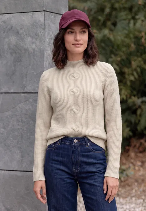 Trui met col en vlechtmotief oat milk beige melange Vrouw met rode pet, beige trui en blauwe jeans voor een stenen muur in de buitenlucht.