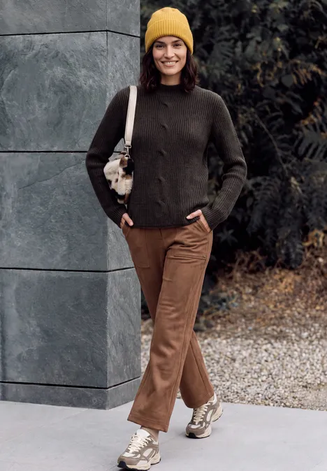 Trui met col en vlechtpatroon truffle brown melange Frau mit gelber Mütze, dunkelgrünem Pullover, braunen Hosen und Sneakern spaziert vor Mauer und Büschen.