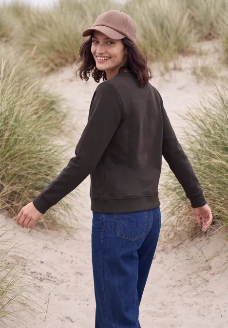 Eine lächelnde Frau in Jeans, brauner Jacke und Cap spaziert am Dünenstrand zwischen Gräsern.