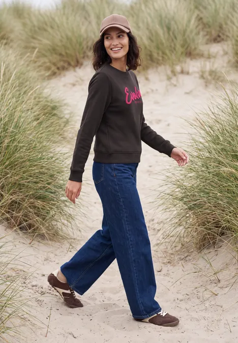 Eine Person spaziert über Sanddünen mit Dünengras, trägt Jeans, Sweatshirt, Mütze und braune Schuhe.