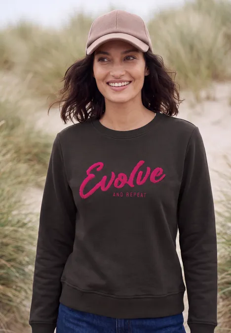 Frau in grauem Sweatshirt mit rotem 'Evolve'-Schriftzug an einem Dünenstrand.