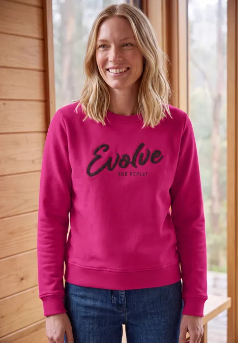 Sweatshirt mit Rundhals und Wording pink flash Eine lächelnde Frau im pinken Pullover mit 'Evolve' Aufschrift vor holziger Fensterkulisse.