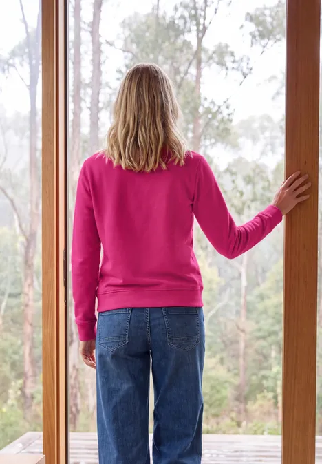 Sweatshirt mit Rundhals und Wording pink flash Person steht mit pinkem Pullover und Jeans im Türrahmen und blickt auf den Wald.