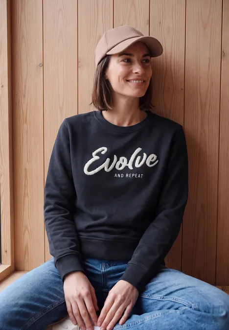Eine Frau sitzt entspannt vor einer Holzwand, trägt Cap, schwarzen Pullover mit Schriftzug und Jeans.