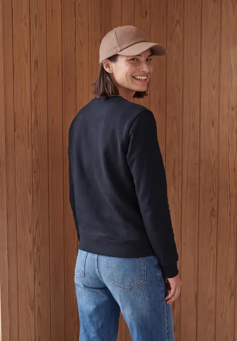 Eine Frau mit Basecap, schwarzem Pullover und Jeans steht vor Holzpaneelen, blickt lächelnd über die Schulter.
