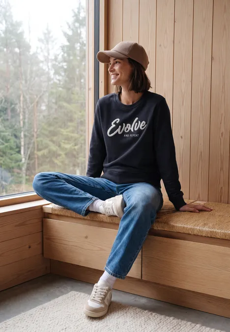 Frau sitzt gemütlich auf Fensterbank in Holzhütte, trägt Jeans, Sneaker und Cap, Pullover mit „Evolve“-Schriftzug.