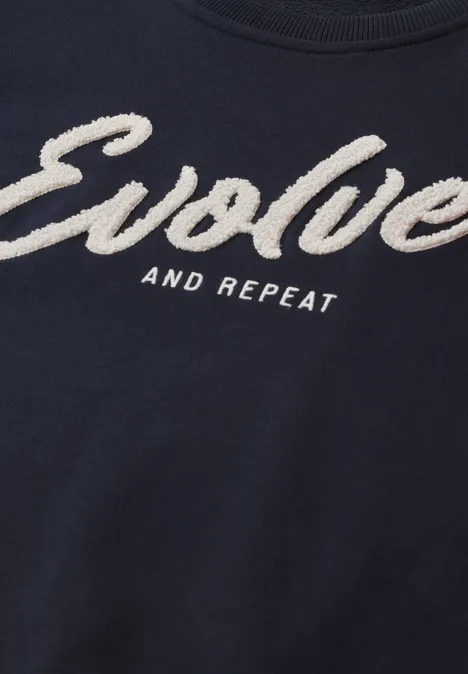 Dunkelblaues T-Shirt mit weißem Schriftzug "Evolve and Repeat" in auffälliger, flauschiger Schrift.