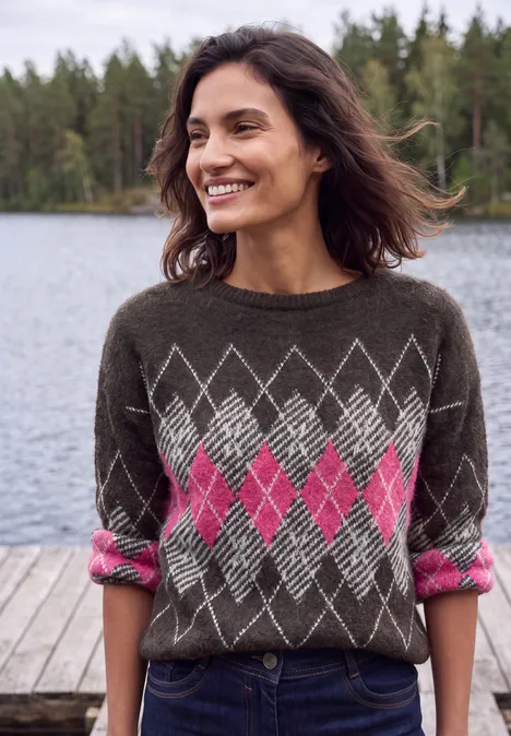 Eine lächelnde Frau in einem braunen Pullover mit pinkem Rautenmuster steht auf einem Steg vor einem See.
