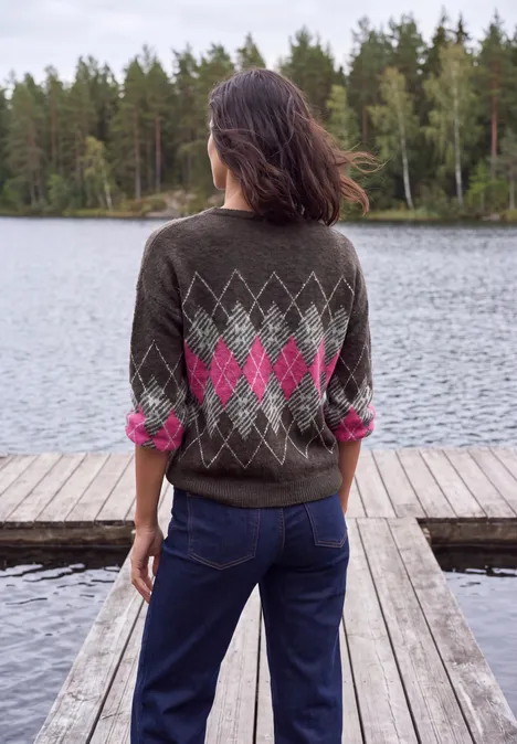 Person im grauen Argyle-Pullover und Jeans steht auf Holzsteg und blickt auf einen von Bäumen umgebenen See.