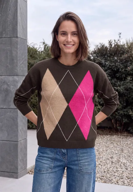 Frau steht lächelnd vor einer Säule im Garten, trägt braunen Pullover mit bunten Rautenmuster und Jeans.