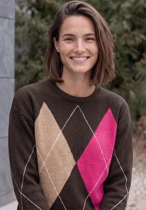 Lächelnde Frau mit kurzen braunen Haaren im braunen Argyle-Pullover mit beigen und pinken Rauten vor Büschen.