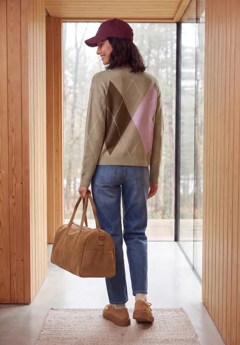 Eine Person in Mütze, gemustertem Pullover, Jeans und Hausschuhen trägt eine Reisetasche in einem modernen Holzhaus.