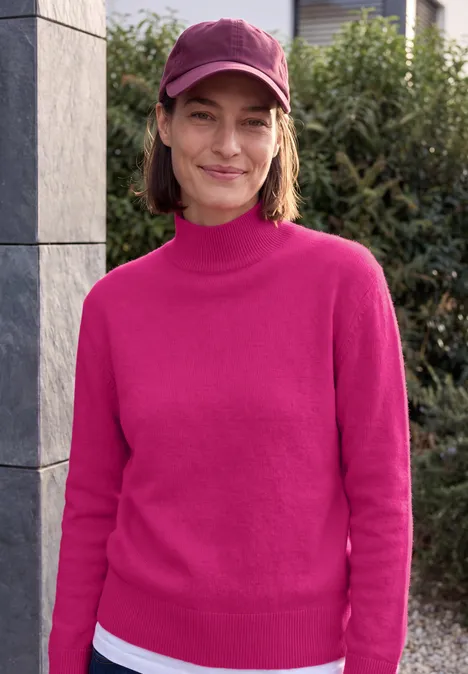 Een vrouw in een roze trui en pet lacht voor een huismuur met planten.