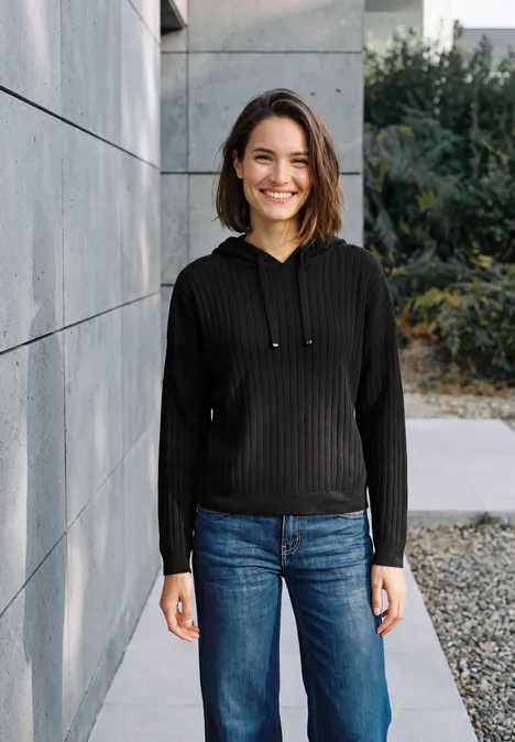 Eine junge Frau in Hoodie und Jeans lehnt an einer Mauer und lächelt in die Kamera.