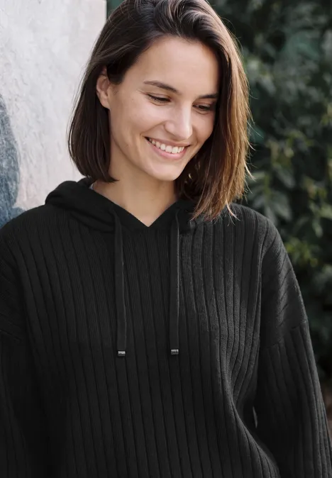 Eine lächelnde junge Frau in schwarzem Pullover mit Kapuze vor grünem Hintergrund.