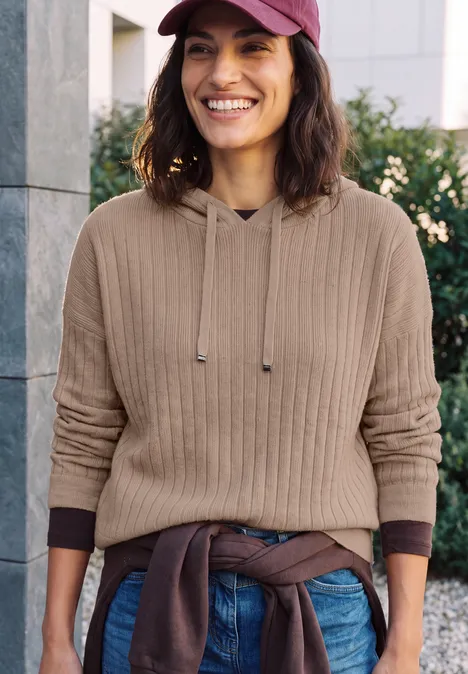 Femme souriante en pull beige, jean et casquette devant un mur urbain.