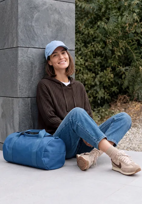 Une femme souriante est assise dehors près d'un mur, porte une casquette et un sac de voyage.