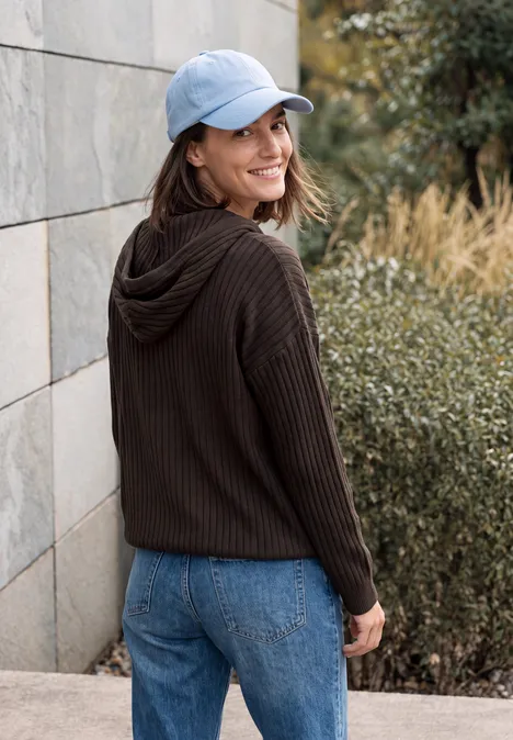 Une jeune femme en jean, sweat à capuche et casquette, adossée à un mur en pierre, souriante en se retournant.