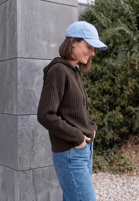 Une femme appuyée contre un mur gris, vêtue d’un sweat à capuche marron, d’un jean et d’une casquette bleu clair.