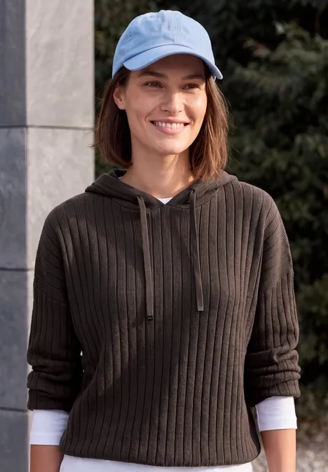 Une femme souriante porte un sweat à capuche côtelé gris foncé et une casquette de baseball bleu clair.