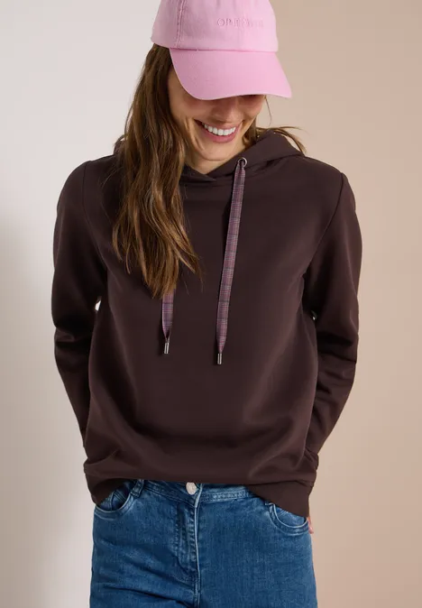 Sweatshirt in Unifarbe mit Kapuze truffle brown Junge Frau lächelt, trägt braunen Kapuzenpullover, blaue Jeans und rosa Baseballcap.