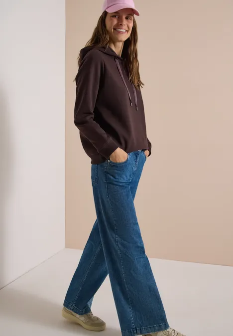 Sweatshirt in Unifarbe mit Kapuze truffle brown Frau mit rosa Cap, braunem Hoodie und weiten Jeans lehnt locker an Wand und lächelt.