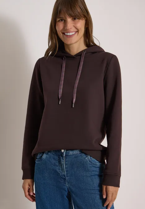 Sweatshirt in Unifarbe mit Kapuze truffle brown Eine lächelnde Frau trägt einen dunkelbraunen Kapuzenpullover und Jeans vor hellem Hintergrund.