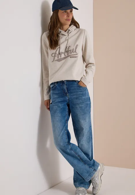 Sweatshirt mit Kapuze und Artwork light cashmere beige melange Frau in beiger Kapuzenjacke, Jeans und Cap lehnt lässig an weißer Wand.