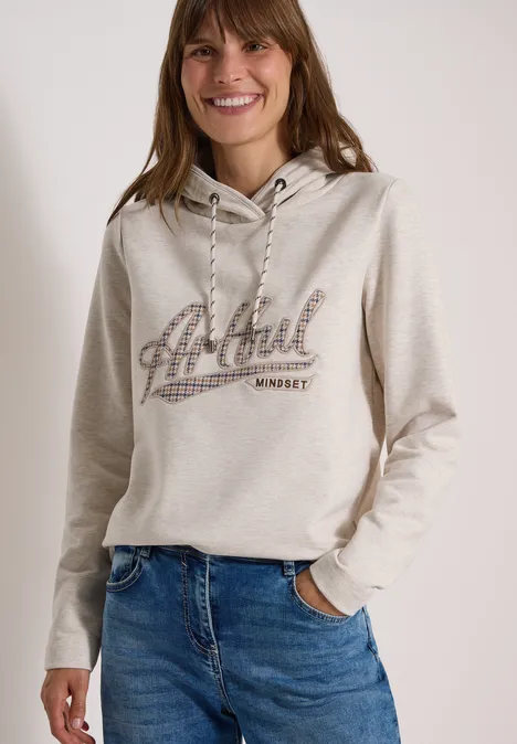 Sweatshirt mit Kapuze und Artwork light cashmere beige melange Lächelnde Frau in beigem Hoodie mit Aufdruck und Jeans, eine Hand in der Tasche.