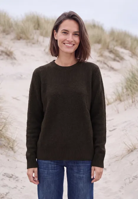 Een vrouw in een donkere trui en spijkerbroek lacht voor grasrijke duinen.