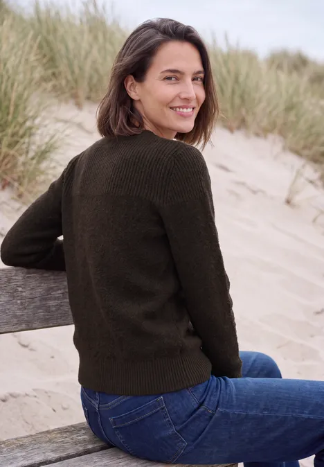 Een glimlachende vrouw in een groen trui en spijkerbroek zit op een bankje aan het strand, duinen op de achtergrond.