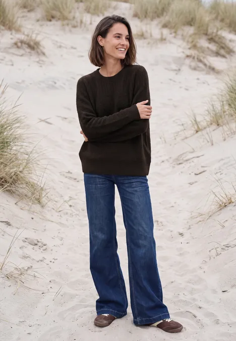 Vrouw lacht op het strand in een donkere trui, wijde blauwe jeans en bruine schoenen voor duinen.