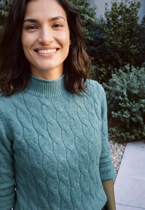 Une femme sourit en extérieur, vêtue d’un pull torsadé vert, devant des arbustes.