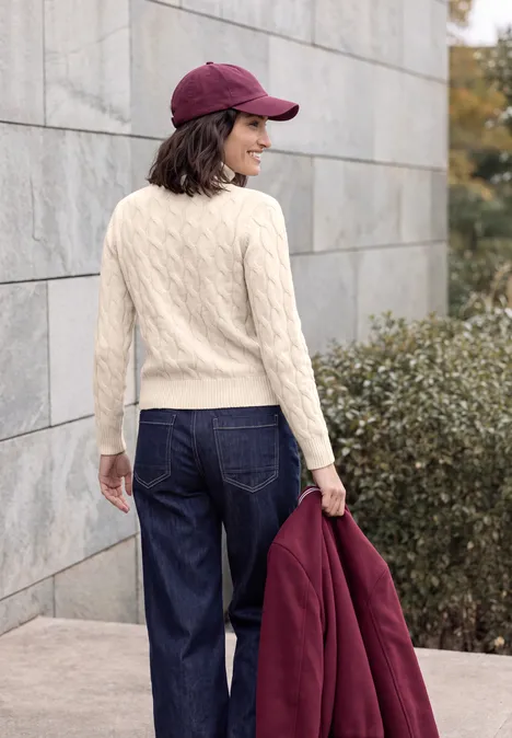 Pullover mit Turtle Neck und Zopfmuster oat milk beige melange Frau in hellem Strickpullover, Jeans und Baseballcap hält eine Jacke und steht vor einer Steinmauer.