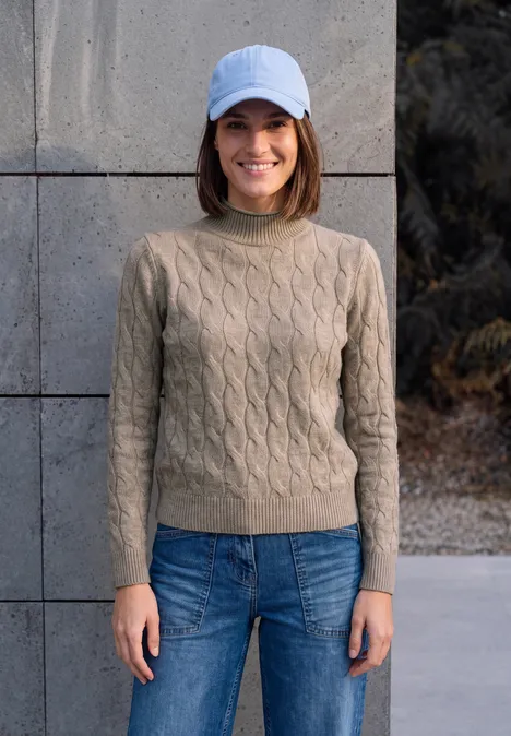 Eine Frau mit weißer Mütze, beigem Strickpullover und Jeans lächelt vor einer grauen Wand.