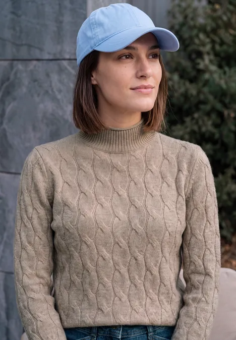 Junge Frau mit hellblauer Baseballkappe und olivgrünem Zopfmuster-Pullover steht draußen vor einer Steinmauer.