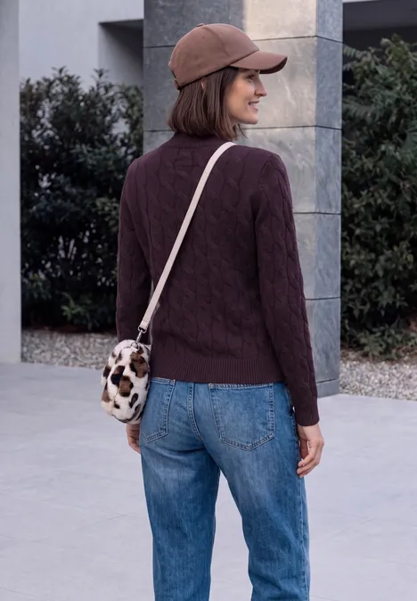 Femme en pull marron, jean bleu, casquette et sac bandoulière motif vache, debout devant un bâtiment.