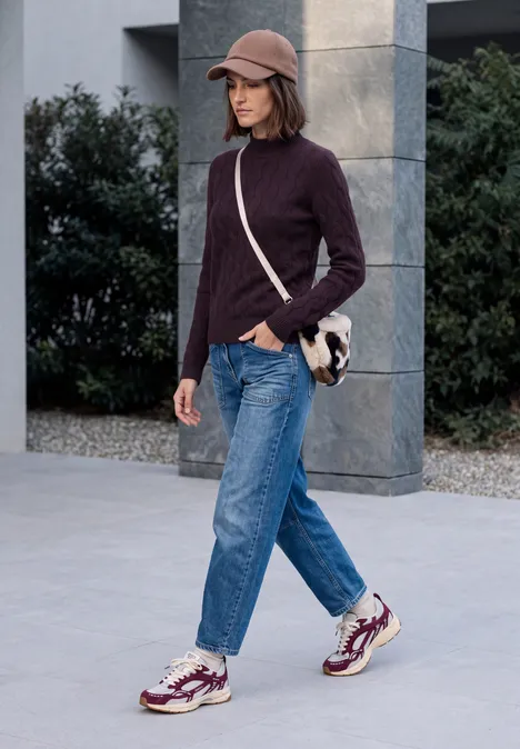 Femme en pull col roulé violet, jean et baskets marche avec sac à bandoulière devant un bâtiment moderne.