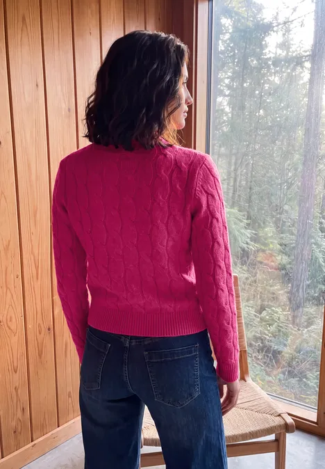 Vrouw in een roze trui en spijkerbroek, gezien van achteren, staand voor een raam tegen een houten wand.