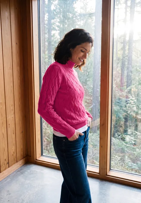 Vrouw in roze trui en spijkerbroek lacht bij een raam met uitzicht op het bos.