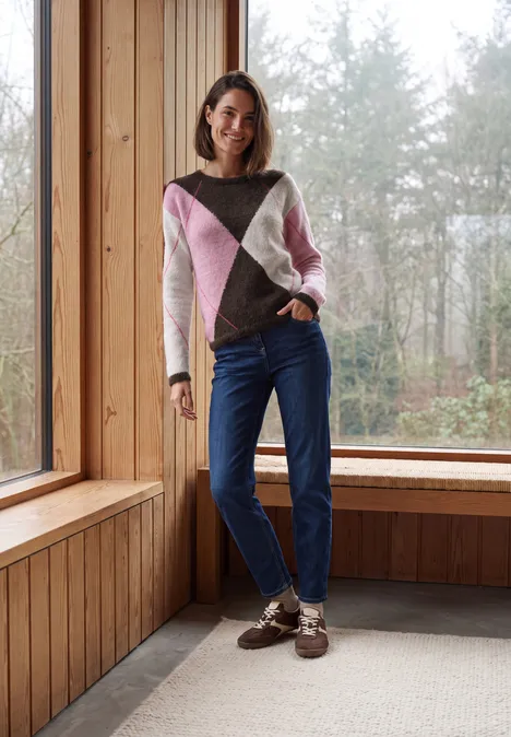 Pullover mit Rundhals und Argyle-Muster truffle brown Frau steht in gemütlichem Holzraum vor großem Fenster mit Wald, trägt Pullover und Jeans.