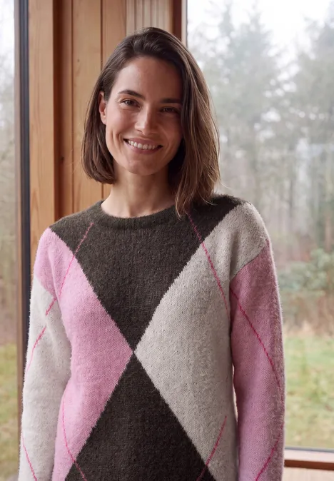 Pullover mit Rundhals und Argyle-Muster truffle brown Frau mit schulterlangem Haar im rosa-weißen Rautenmuster-Pullover lächelt vor einem Fenster mit Waldblick.